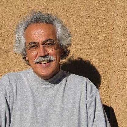 An older man with gray curly hair, glasses, and a mustache, wearing a light gray turtleneck sweater, smiles in front of a plain tan-colored wall in sunlight.