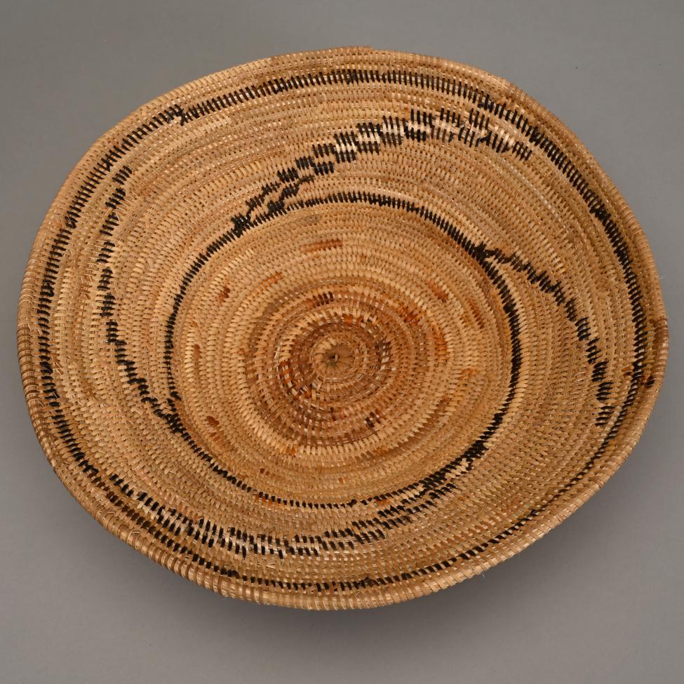 A round woven basket with a spiral pattern in natural tan colors, featuring accents of darker brown and black, displayed on a plain gray background.
