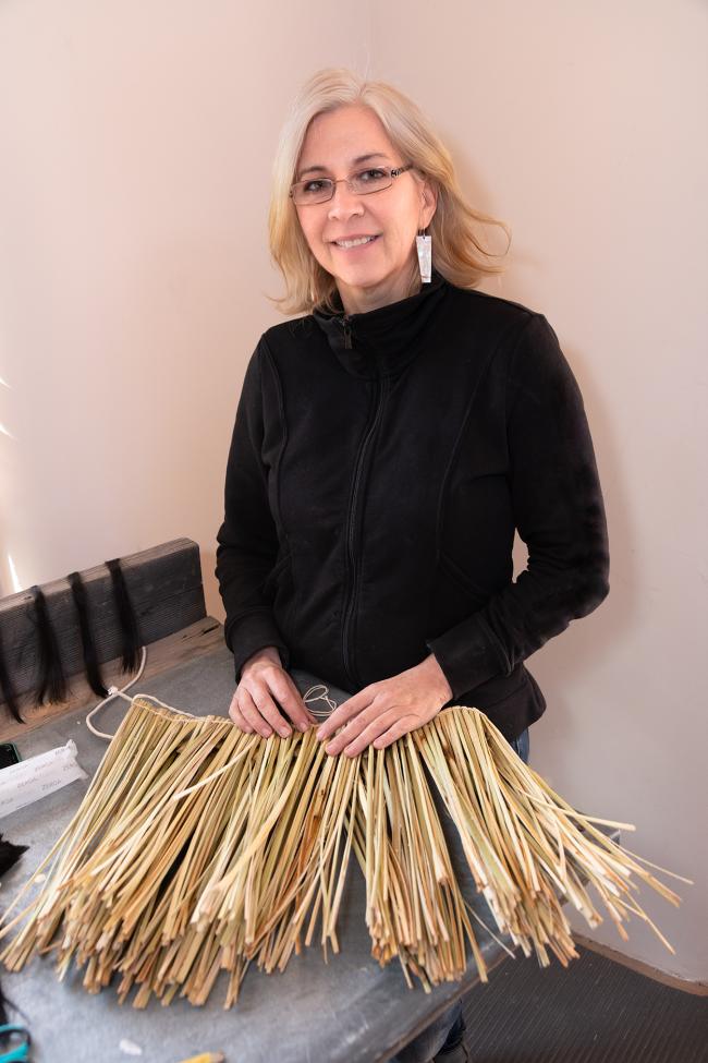 A woman with light hair and glasses, wearing a black jacket, stands smiling at a table covered with bundles of long, thin reeds or straw, likely used for weaving or crafts.