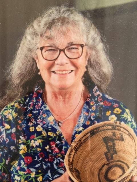 Smiling older woman with gray hair and glasses, wearing a floral shirt, holds a woven basket with a geometric design.
