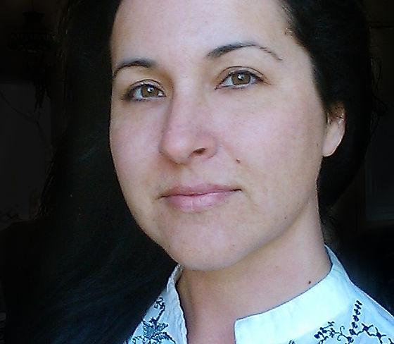 A woman with long dark hair and light skin looks at the camera with a soft expression. She is wearing a white top with black embroidery. The background is dark and out of focus.