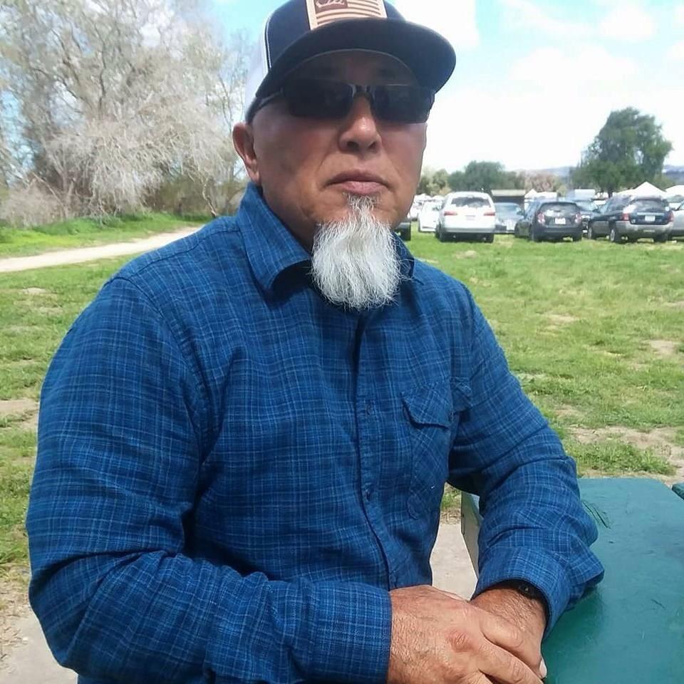 A man with a gray goatee, sunglasses, and a dark cap sits outdoors at a green table, wearing a blue plaid shirt. There are trees, grass, and parked cars in the background on a sunny day.