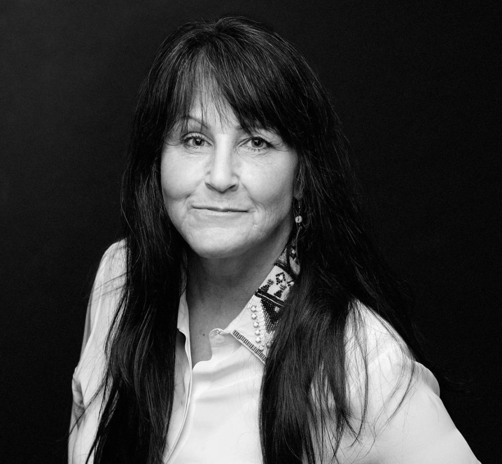 Black and white portrait of a middle-aged woman with long dark hair, wearing a light-colored shirt with a patterned collar, looking at the camera and smiling slightly, against a plain dark background.