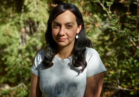 A woman with long dark hair, wearing a light-colored t-shirt with illustrated figures, stands outdoors in sunlight, with green foliage and tree branches in the background.
