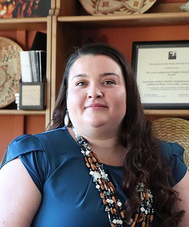 A woman with long brown hair wearing a blue top and a beaded necklace smiles softly. She stands in front of shelves displaying woven baskets, a framed certificate, and decorative items.