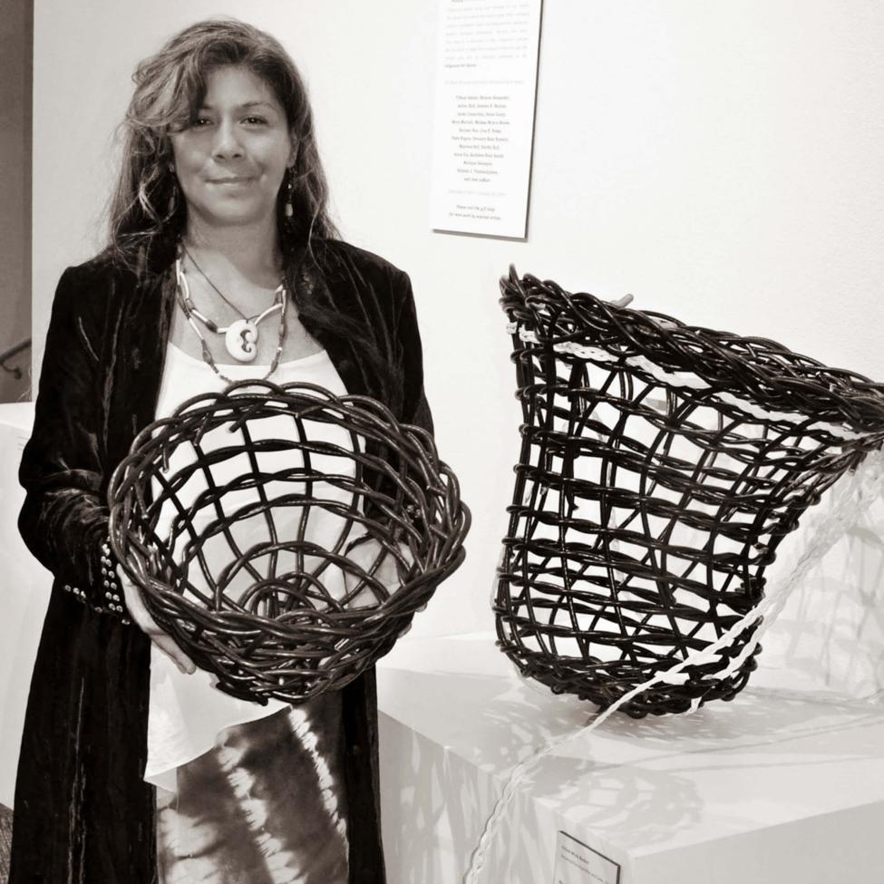A woman with long hair holds a woven basket and stands beside another similar basket displayed on a pedestal in an art gallery. She is dressed in a tie-dye skirt, white top, dark jacket, and a large beaded necklace.