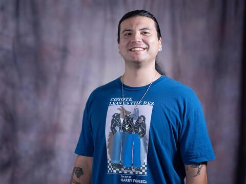 A smiling man with long dark hair, wearing a blue T-shirt with artwork and text on the front, stands in front of a blurred, purple-gray backdrop. He has tattoos on his arms and is wearing earrings and a necklace.