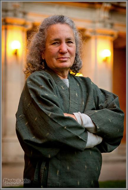 An older woman with curly gray hair stands outdoors with her arms crossed, wearing a green robe-like garment. Warm lights and a stone building are visible in the background.