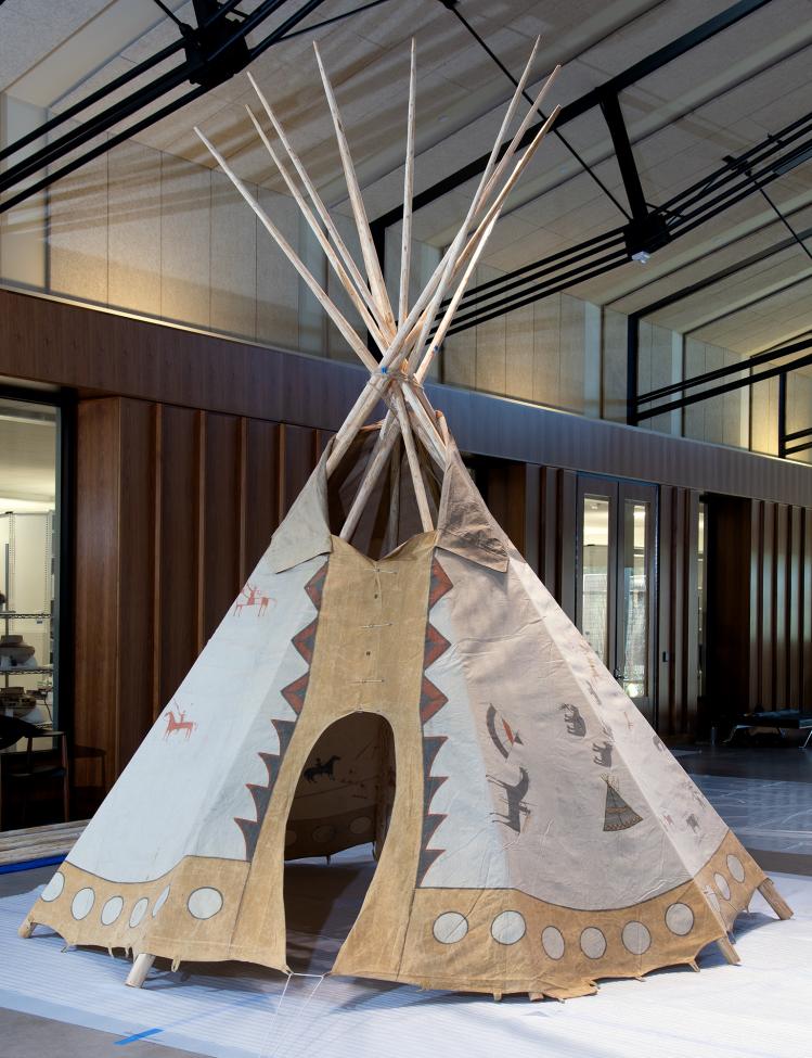 A traditional tipi with painted designs stands indoors on a modern floor, surrounded by wood-paneled walls and large windows in a spacious, contemporary building.