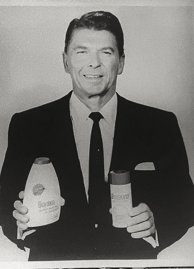 A man in a suit and tie smiles while holding two containers of Boraxo hand soap, one in each hand, against a plain background. The image is in black and white.
