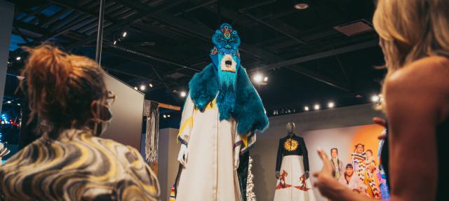 Two people observe an elaborate costume on display in a dimly lit exhibition. The costume features vibrant blue fur and an intricate mask with a headdress. Another costume on a mannequin and various colorful outfits are visible in the background.