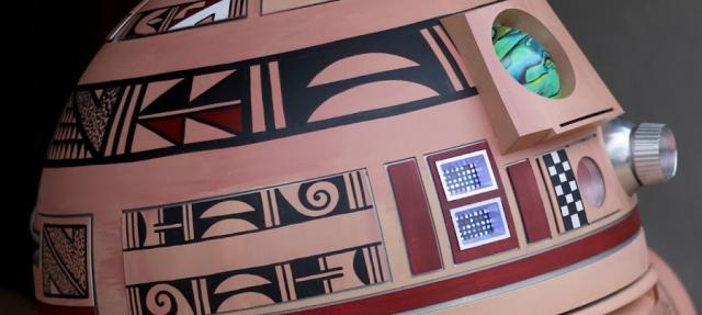 A robot resembling a cultural design with geometric patterns and earthy colors. Its cylindrical body features intricate motifs in red, brown, black, and white. The head has a circular green sensor and metallic details.