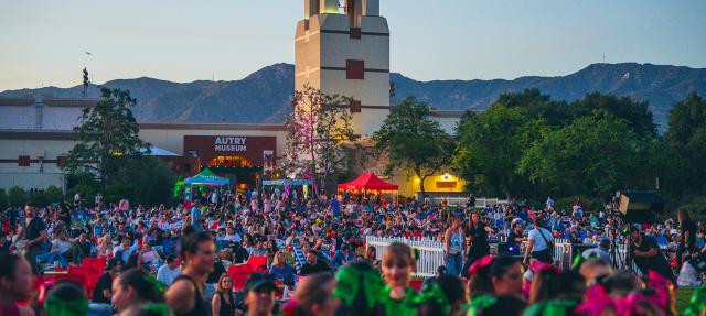 A large crowd gathers outdoors in front of the Autry Museum at dusk, with mountains in the background and a tower visible. People sit on chairs and blankets; festive decorations and tents add to the lively atmosphere.