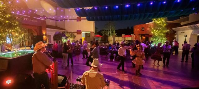People in western attire dance under string lights at an outdoor evening event at the Autry Museum. Musicians perform on stage in the foreground; trees and buildings are decorated with festive lights.