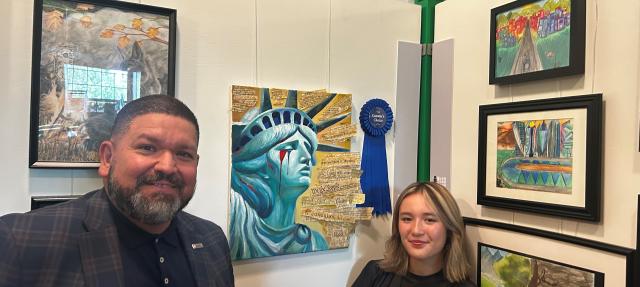 A man and a young woman stand smiling in front of a display of colorful student artwork, including a painting of the Statue of Liberty with a blue ribbon attached, at what appears to be an art show or school event.