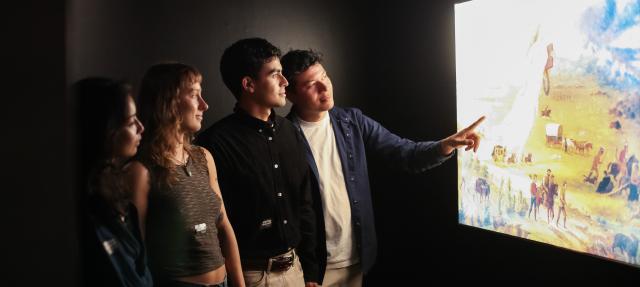 Four people stand in a dark room closely observing and discussing a brightly lit display or painting on the wall, with one person pointing at the artwork.