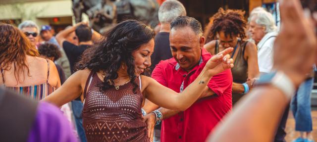 A woman and man dance joyfully outdoors in a crowd, surrounded by people smiling and enjoying the lively atmosphere. The woman raises her arm while the man smiles, closely following her moves.