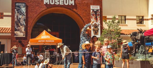 A group of children and adults enjoy a lasso activity in front of the Autry Museum, with people wearing cowboy hats and a bright orange museum tent visible on a sunny day.