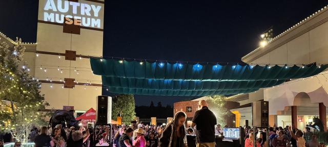 A large crowd attends an outdoor nighttime event at the Autry Museum, with string lights overhead, people mingling, and a DJ booth in the foreground. The museum’s name is projected on a building wall.