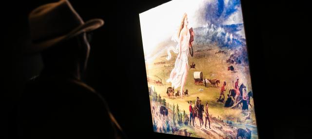 A person in a hat stands in a dark room, looking at an illuminated painting depicting pioneers, covered wagons, and a large female figure in flowing white robes over a landscape.