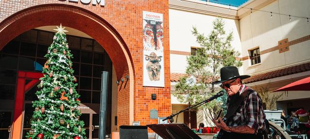 A musician in a cowboy hat plays guitar and sings outside the Autry Museum, near a large decorated Christmas tree. The scene is sunny, with festive holiday decorations and museum banners visible.