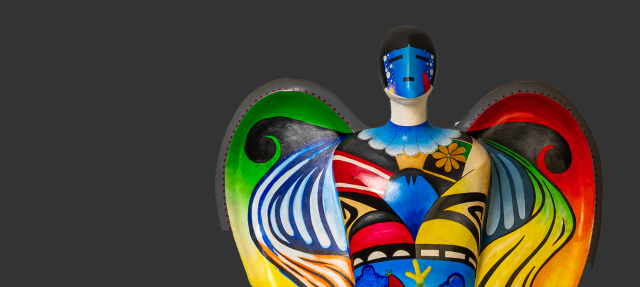 A colorful, abstract angel sculpture with large wings, decorated in vibrant blue, green, red, yellow, and black patterns, stands against a plain dark gray background.