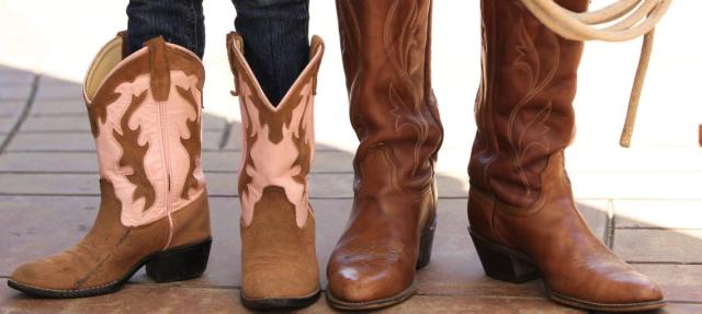 Two people wearing cowboy boots stand side by side on a tiled surface. The pair on the left are shorter with pink and brown designs, while the boots on the right are taller and plain brown. A lasso is held to the right.