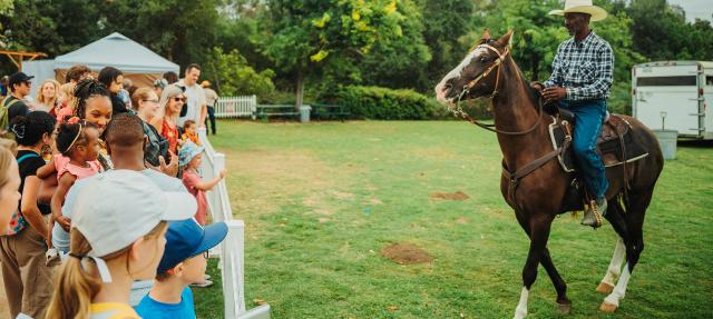 A man wearing a cowboy hat rides a horse near a fenced area where a diverse group of people, including children and adults, watch him outdoors on a grassy field with trees in the background.