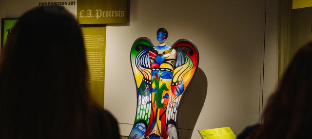 A colorful angel sculpture with abstract patterns is displayed in a museum exhibit about L.A. protests, with two people viewing it from the foreground.