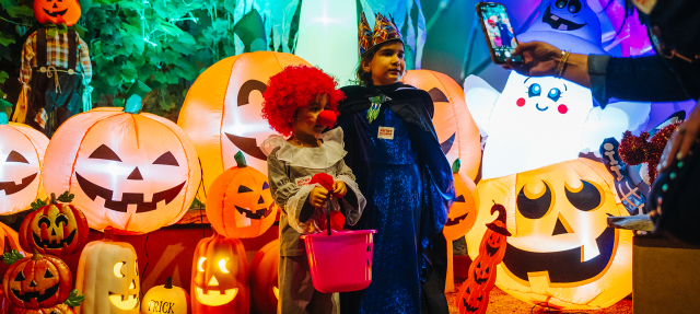 Two children in Halloween costumes stand among large glowing jack-o’-lantern decorations while someone takes their photo. The scene is colorful and festive with spooky, playful decorations.