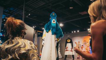 Two people observe an elaborate costume on display in a dimly lit exhibition. The costume features vibrant blue fur and an intricate mask with a headdress. Another costume on a mannequin and various colorful outfits are visible in the background.
