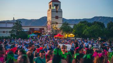 A large crowd gathers outdoors in front of the Autry Museum at dusk, with mountains in the background and a tower visible. People sit on chairs and blankets; festive decorations and tents add to the lively atmosphere.