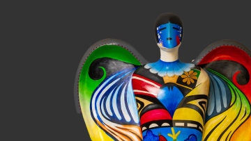 A colorful, abstract angel sculpture with large wings, decorated in vibrant blue, green, red, yellow, and black patterns, stands against a plain dark gray background.