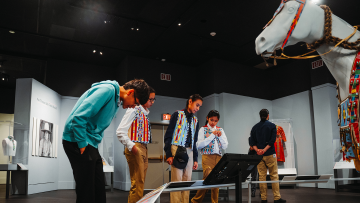 Several people observe exhibits in a museum, including a white horse statue decorated with colorful beadwork. The room has art displays and black-and-white photographs on the walls.