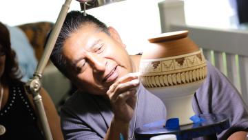 A man smiles while carefully painting intricate designs on a ceramic pot under a desk lamp, focusing on his detailed artwork.