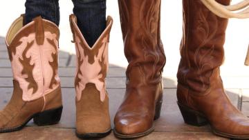 Two people wearing cowboy boots stand side by side on a tiled surface. The pair on the left are shorter with pink and brown designs, while the boots on the right are taller and plain brown. A lasso is held to the right.