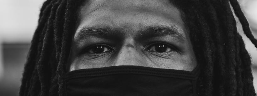 black and white closeup photo of a Black individual with dreadlocks and a mask covering the lower half of their face, leaving their eyes visible to look at the camera