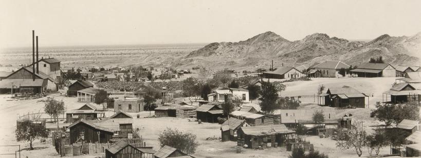 black and white photograph of a small town in the desert, with dirt hills in the background of the buildings