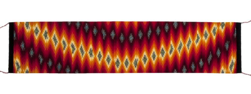 A long, rectangular textile with a vibrant zigzag pattern featuring alternating colors of red, yellow, orange, and black. The design is flanked by black borders on the shorter sides, each with a small red tassel at the end.