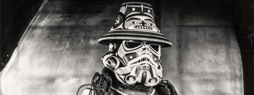 A person dressed in an intricately designed costume featuring Native American patterns and symbols, including a mask resembling a Stormtrooper helmet and a wide-brimmed hat. They hold a small decorated object in one hand and wear multiple necklaces. Black and white photo.