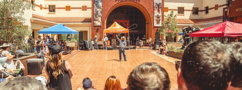 People gathered outdoors watching a performer at the Autry Museum. The museum's entrance is in the background, with banners on either side and tents providing shade over some spectators. The scene is lively with a mix of standing and seated onlookers.