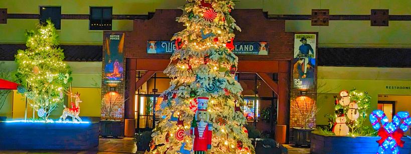 A festive holiday display featuring a large decorated Christmas tree adorned with lights and ornaments, with a nutcracker figure in front. The backdrop includes a building entrance with additional lit decorations, banners, and a snowman made of lights.