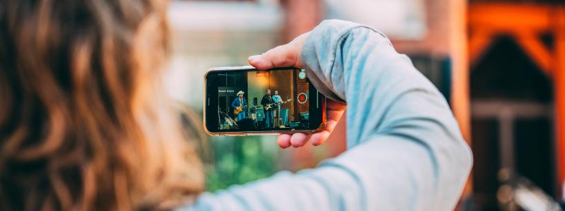 A person with wavy hair and wearing a light gray long-sleeve shirt holds up a smartphone, capturing a photo of a band performing on stage. The stage backdrop features a combination of brick and wooden elements, illuminated with warm lighting.