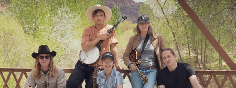 Five people pose with musical instruments on a wooden bridge surrounded by greenery and mountains. From left to right: they hold a violin, banjo, acoustic guitar, mandolin, and another violin. Some wear hats and casual attire, and they appear relaxed and smiling.