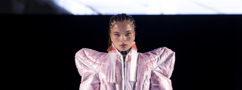 A model walks down a lit runway wearing a metallic pink, futuristic, padded jumpsuit with exaggerated shoulders. Her hair is styled in braids, and she pairs the outfit with white platform shoes and red statement earrings. The background is dark, highlighting the runway.