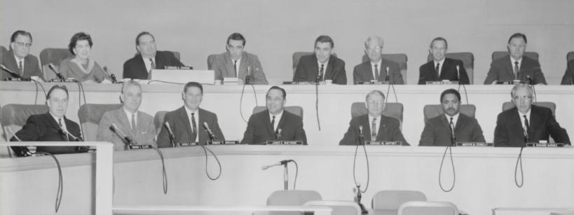 A black-and-white photo shows a group of people seated in a formal setting, likely a conference room or council chamber. They are arranged in two rows behind microphones on desks. The room has a modern design with clean lines and minimal decor.