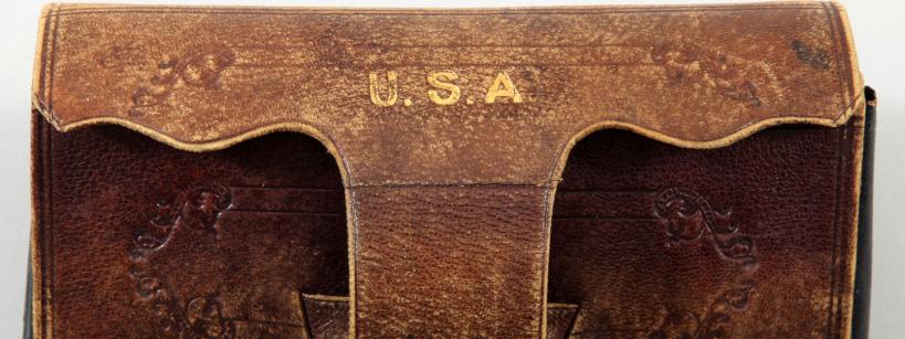 Vintage, well-worn brown leather pouch with intricate embossed designs and the letters "U.S.A" stamped in gold at the top. The pouch appears to have a flap closure and shows signs of aging with a slightly faded texture.