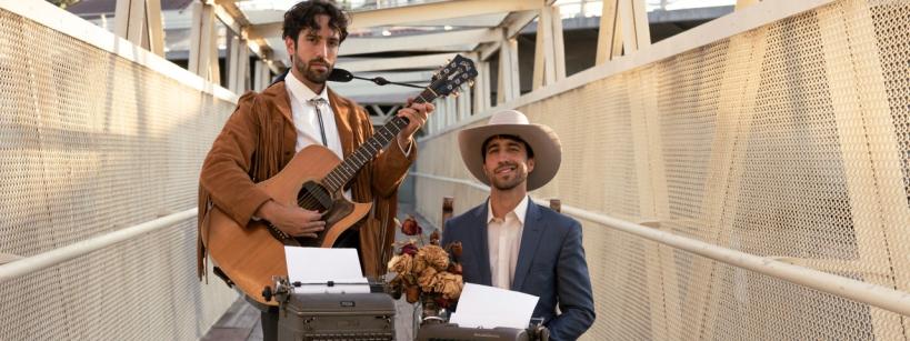 Two men standing on an outdoor bridge with a netted railing. One stands playing an acoustic guitar, wearing a brown suede jacket with fringe. The other sits at a table covered with a floral cloth, wearing a white hat and suit, with two typewriters in front of him.