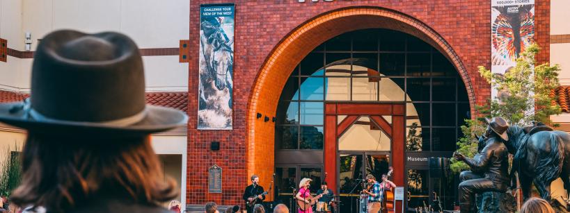 Outdoor concert at the Autry Museum with musicians performing on stage. A crowd watches, including a person in the foreground wearing a wide-brimmed hat. Brick building and statues enhance the vibrant scene.