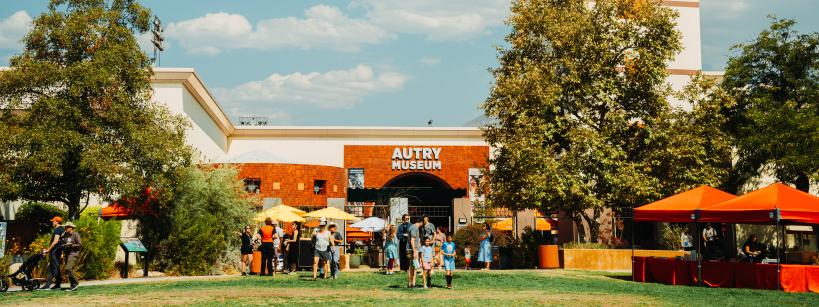 A sunny day at the Autry Museum, featuring a large entrance tower, with people gathered outside. Trees and orange tents are in the foreground, and the sky is clear with a few clouds.